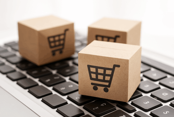 Trzy kartonowe pudełka z symbolem koszyka stojące na klawiaturze laptopa, symbolizujące cyfrową obsługę zamówień w portalu B2B zintegrowanym z systemem ERP.