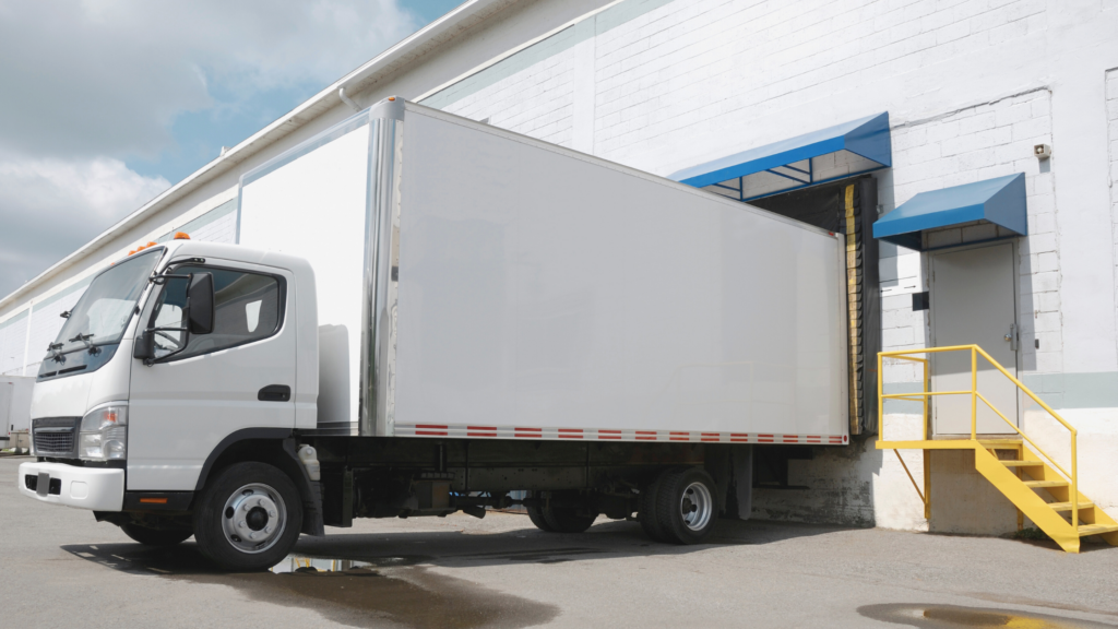 Biała ciężarówka przy rampie rozładunkowej magazynu podczas dostawy towaru.