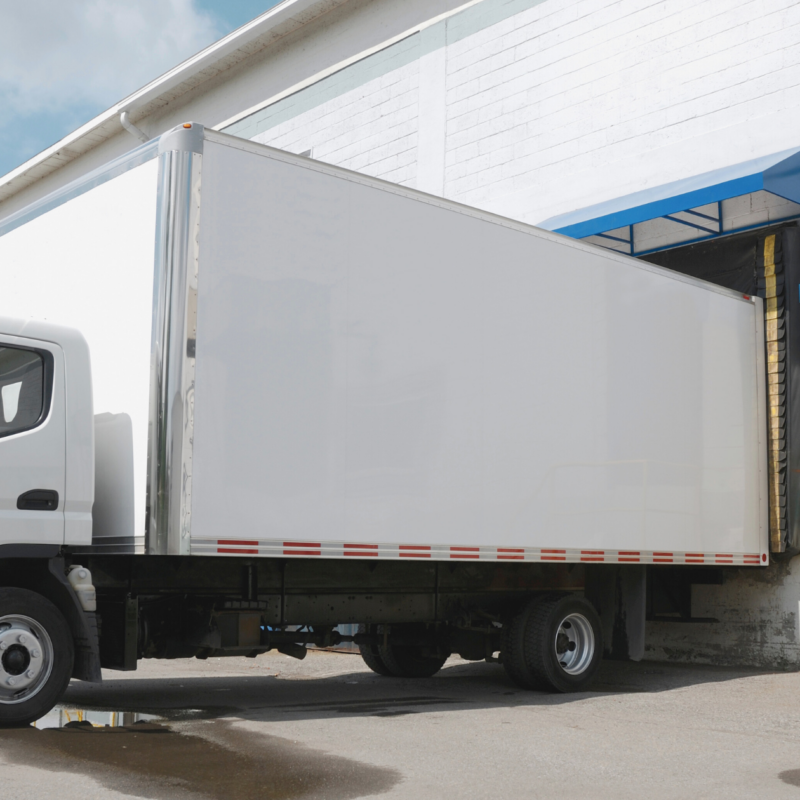 Biała ciężarówka przy rampie rozładunkowej magazynu podczas dostawy towaru.