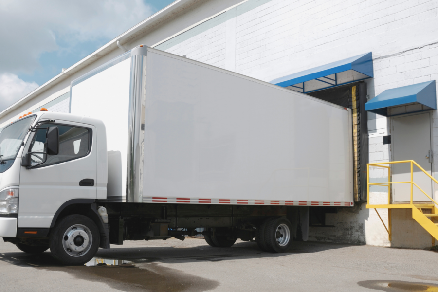Biała ciężarówka przy rampie rozładunkowej magazynu podczas dostawy towaru.